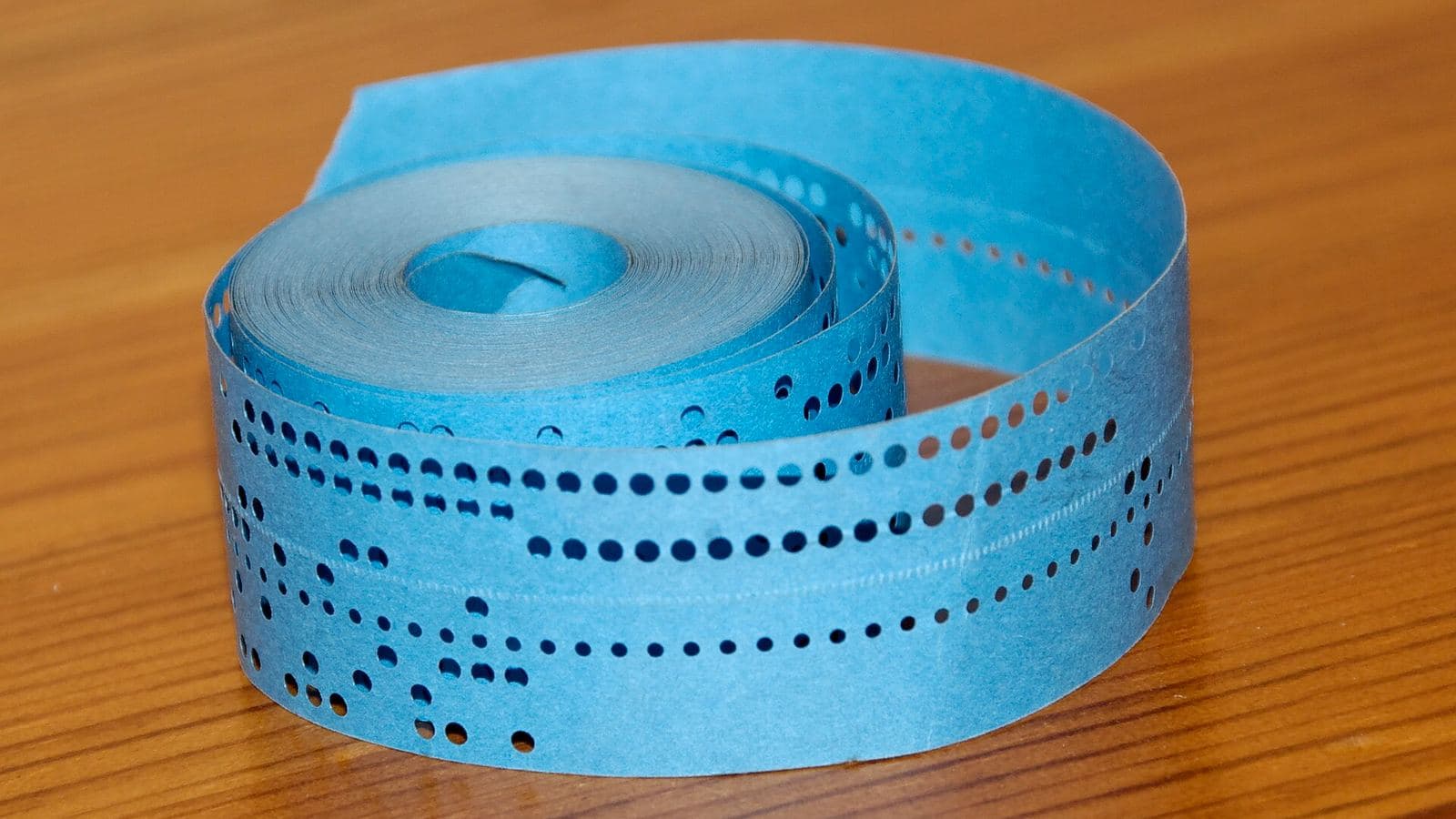 A coil of pale blue punched paper tape standing on a wooden table, its rows of round punched holes catching the light.