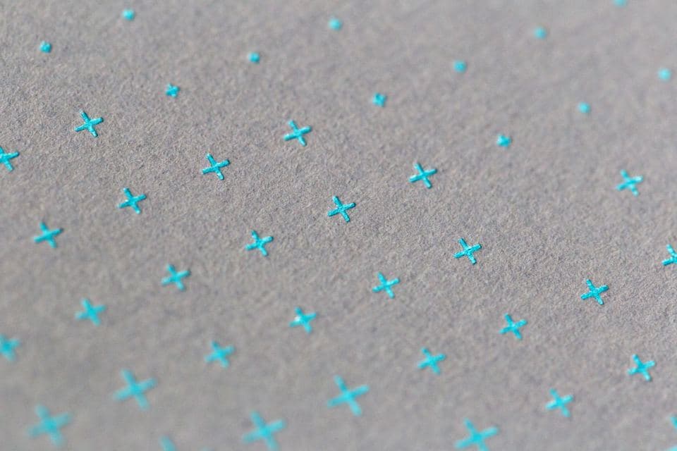 Grey textured paper printed with a grid of small turquoise foil crosses, lit at an angle.