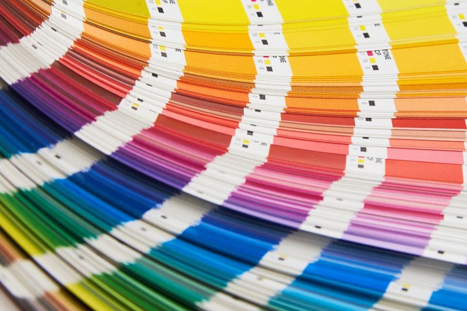 Fanned strips of printed ink colour scales, showing process yellow through oranges, reds, magentas and blues on white card printed with registration marks.