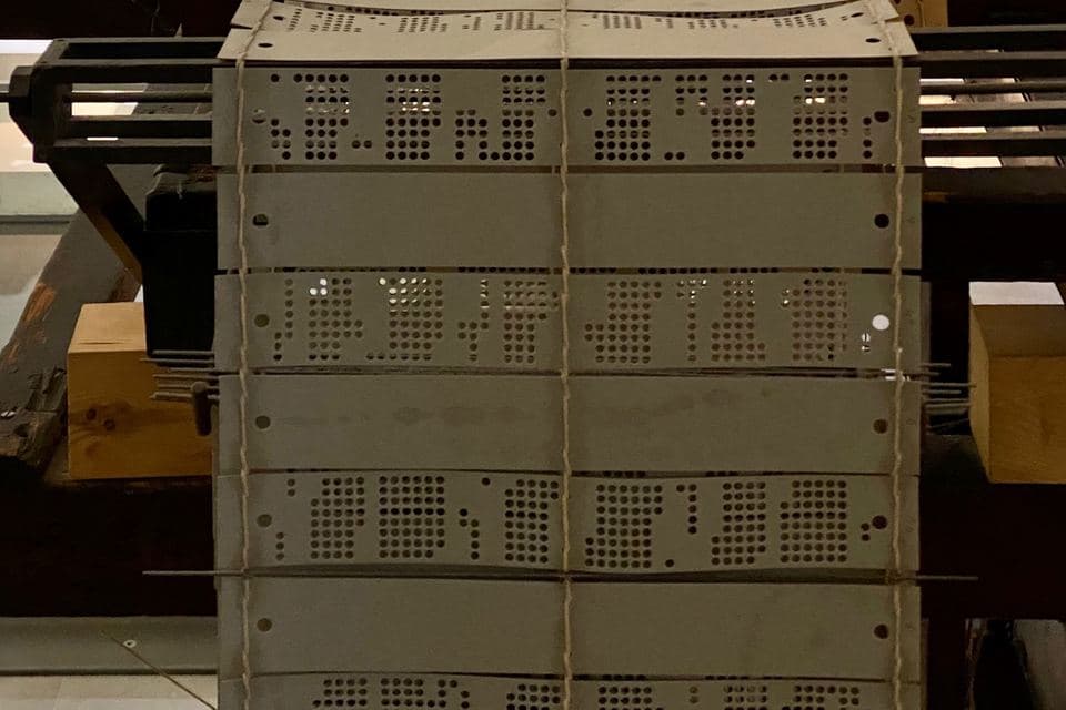 Rows of grey punched cards laced together with string and mounted in a loom, each card filled with grids of punched holes, with wooden machine parts either side.