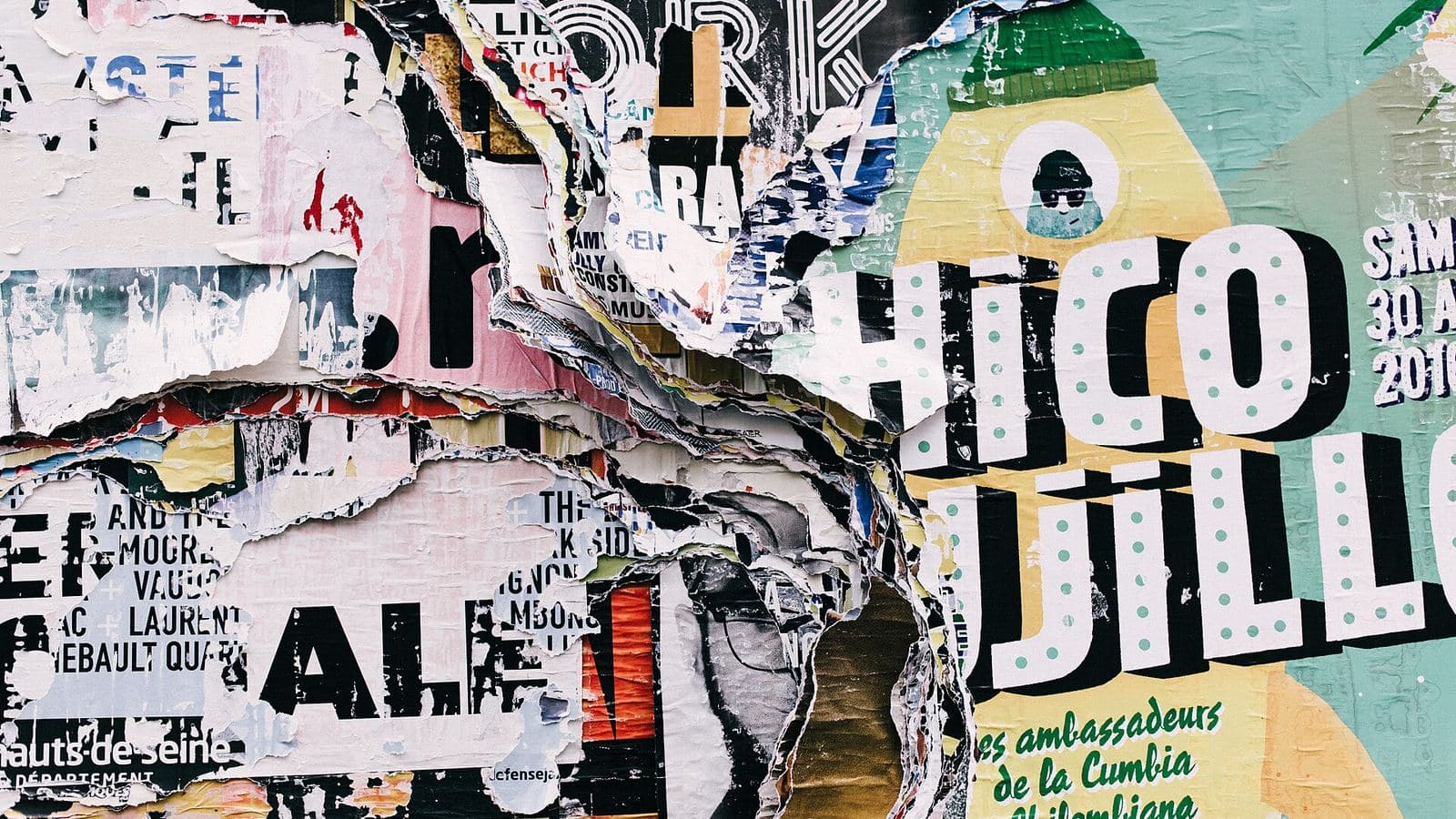 Layers of torn concert posters pasted onto a wall, black-and-white type fragments showing through and a teal and yellow sheet with polka-dot lettering on top.