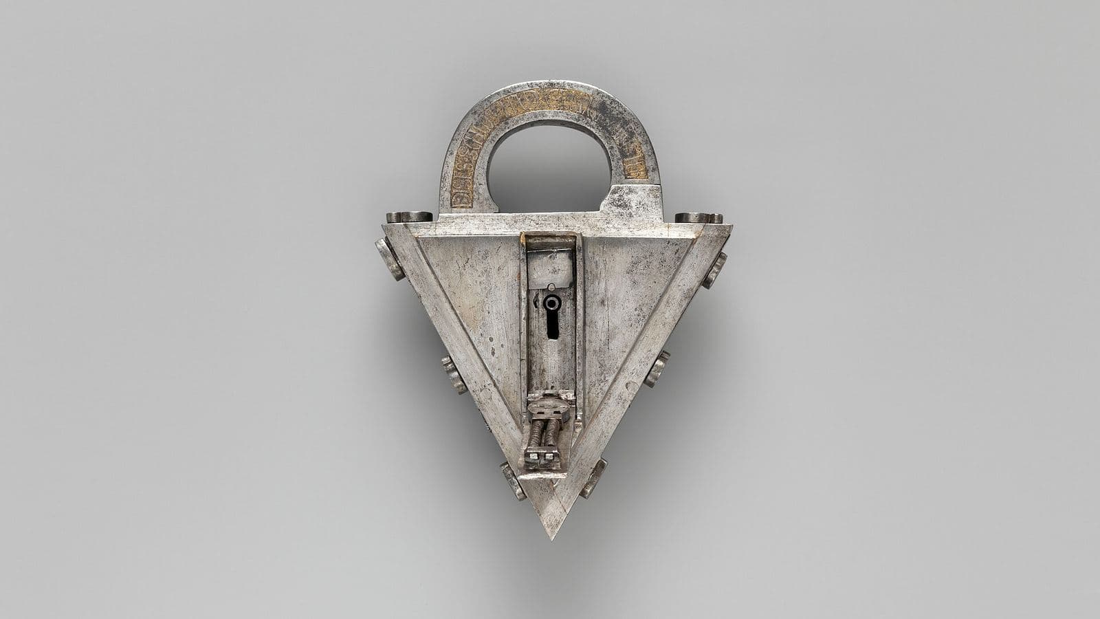 A steel padlock with gilded lettering on its shackle, standing upright against a pale grey ground, its faceplate open on a small exposed spring mechanism.